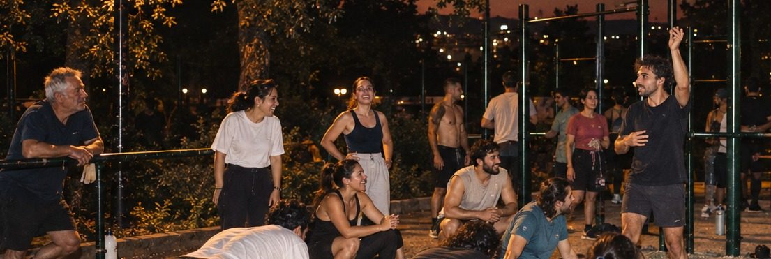 A free community training at night: a coach with a raised arm talks to a group doing push-ups while others hang from the bars and watch, laughing