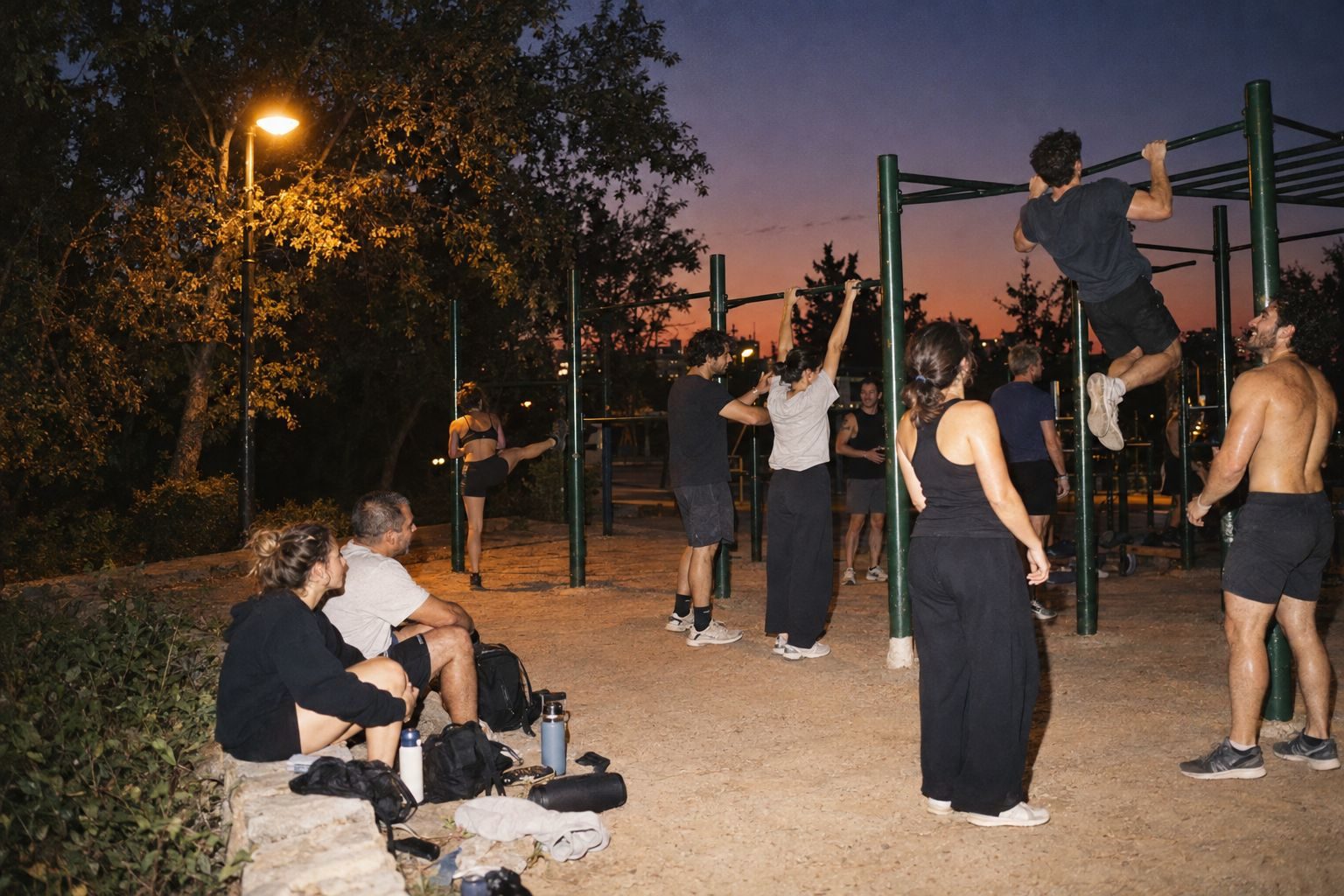 Night at the bars: someone mid pull-up while friends watch, two sit on a low wall with bags and a speaker